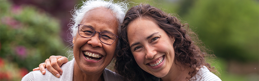 A caregiver and an elderly woman smile together outdoors.